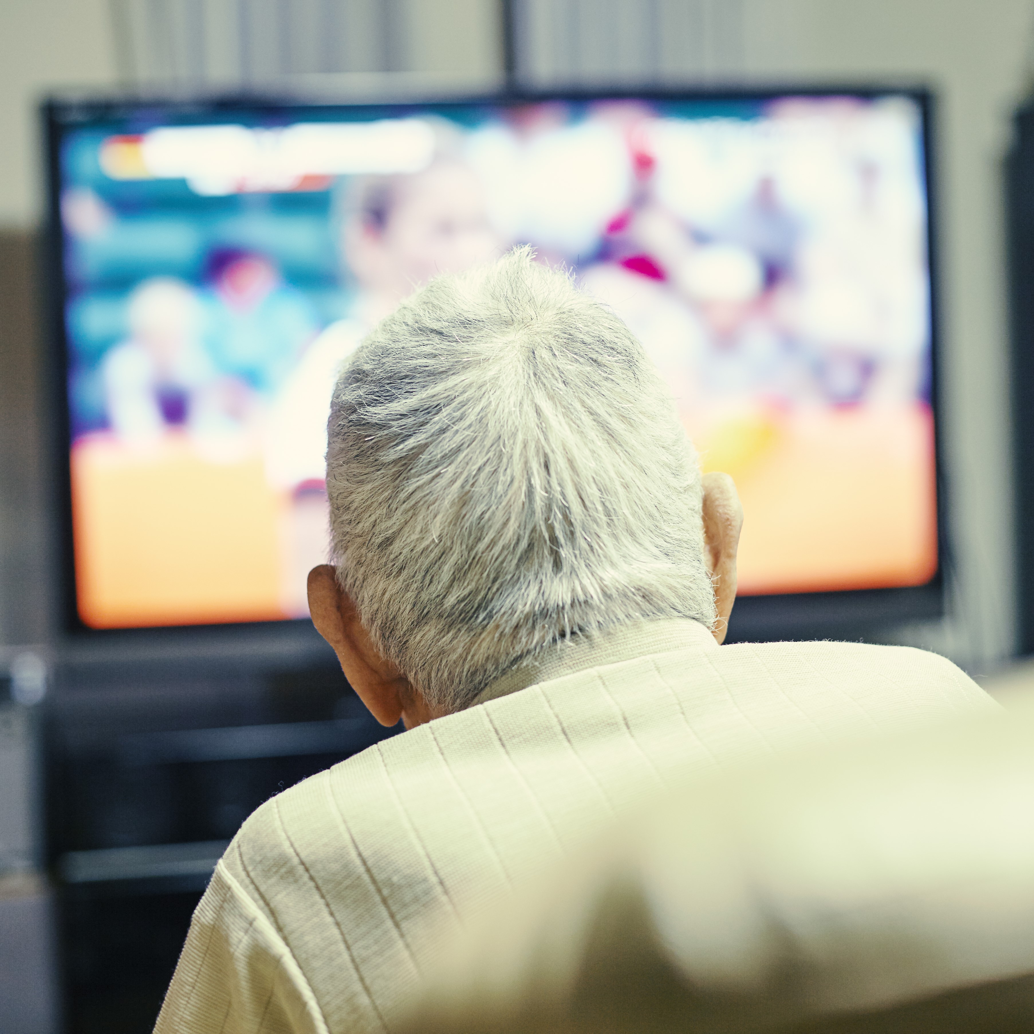 The head and shoulders of a white-haired older man are seen from the back. He is watching television. 